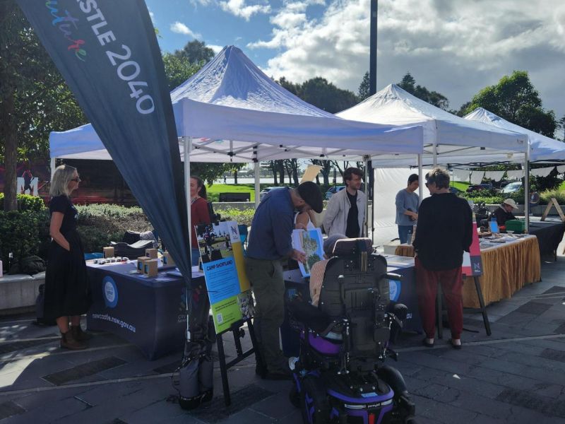 A community member is seen conversing with a City of Newcastle staff member in front of a marquee at The Station. Project corflutes and corflutes are also pictured, with two City of Newcastle staff in conversation with one another in the background.