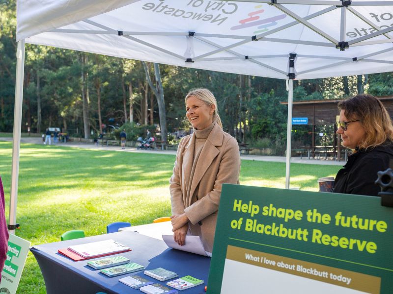 A City of Newcastle staff member is seen under a marquee, conversing with a community member who is not pictured, at Blackbutt Reserve.