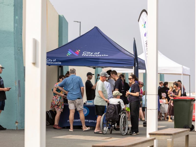 A number of people gather around and in front of a marquee to converse with City of Newcastle staff at Newcastle Ocean Baths.