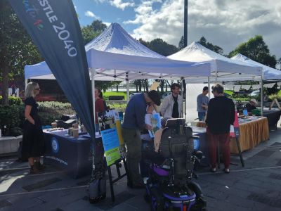 A City of Newcastle marquee set up outdoors at The Station. City of Newcastle staff are seen engaging in conversation with market attendees, in front of project signage..