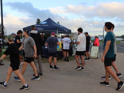 A City of Newcastle marquee set up outdoors at Nobbys Beach at sunrise. City of Newcastle staff are seen engaging in conversation with a local running club and others passing by.